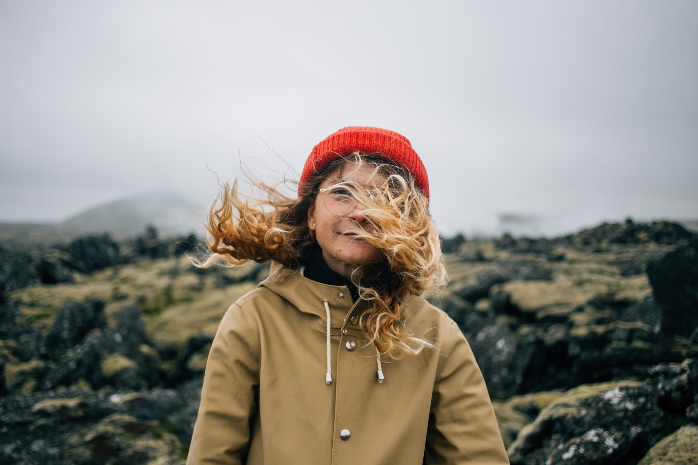Hipster millennial young woman in raincoat and trendy beanie looks into camera. Stand in middle of epic rough iceland landscape, wind blows blond hair in face. romantic inspiration wanderlust Happy young woman smiles to camera in wind