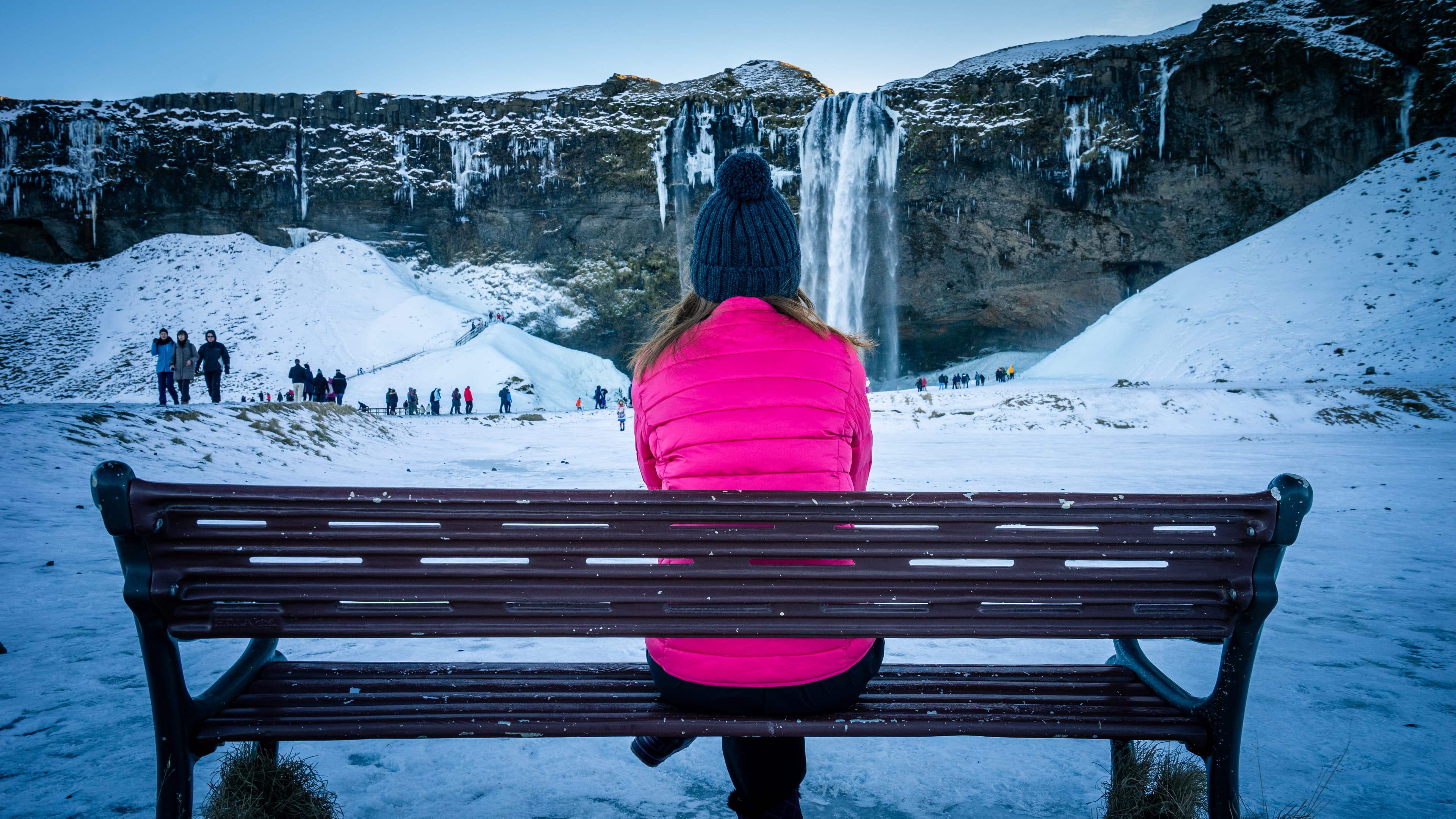 A girl in pink jacket sits on the bench and looking tourists in front of the frozen Seljalandsfoss waterfall in wintertime. It is one of most popular place in South Coast Iceland. A girl in pink jacket sits on the bench and looking tourists in front of the frozen Seljalandsfoss waterfall in wintertime. It is one of most popular place in South Coast Iceland.