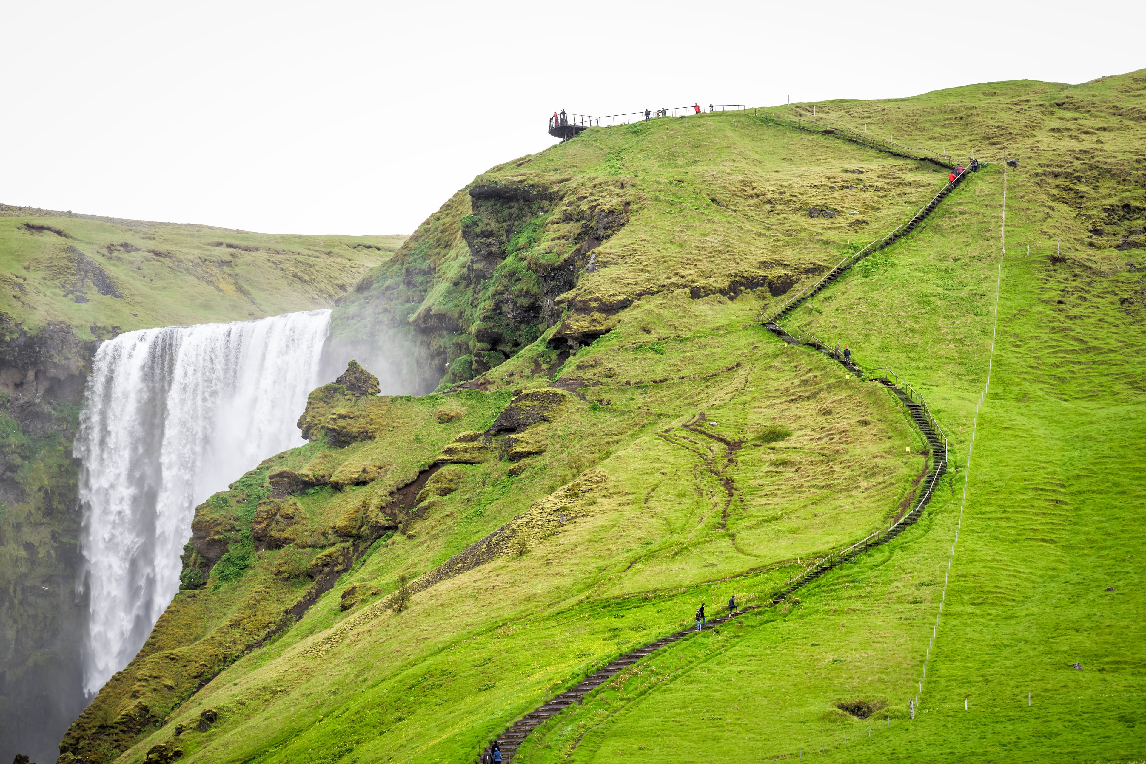 Skogafoss, Iceland Waterfall on cliff with green grass hills and many tourists people walking on steep high trail road in mossy summer Skogafoss, Iceland Waterfall on cliff with green grass hills and many tourists people walking on steep high trail road in mossy summer