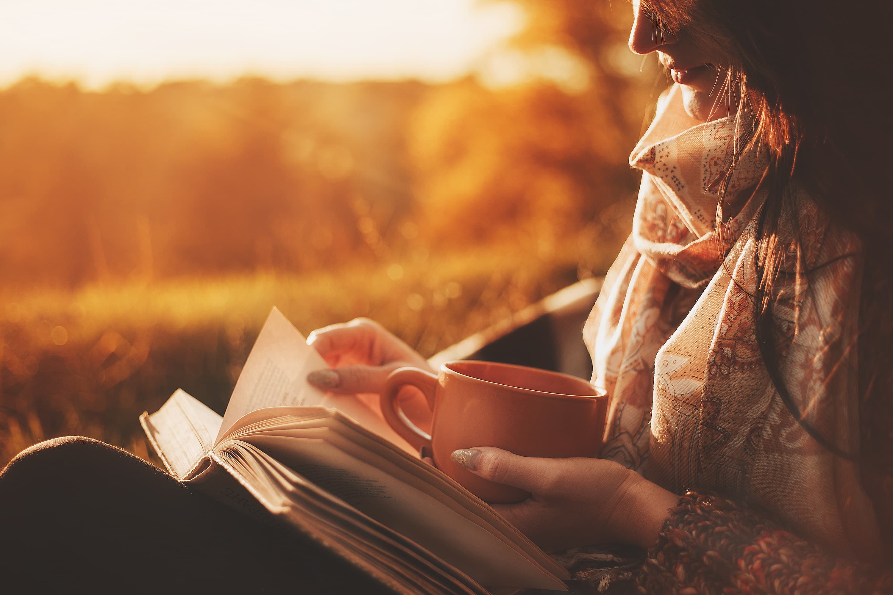 Beautiful girl in autumn forest reading a book covered with a warm blanket.a woman sits near a tree in an autumn forest and holds a book and a cup with a hot drink in her hands. Girl reading a book a woman sits near a tree in an autumn park and holds a book and a cup with a hot drink in her hands. Girl reading a book