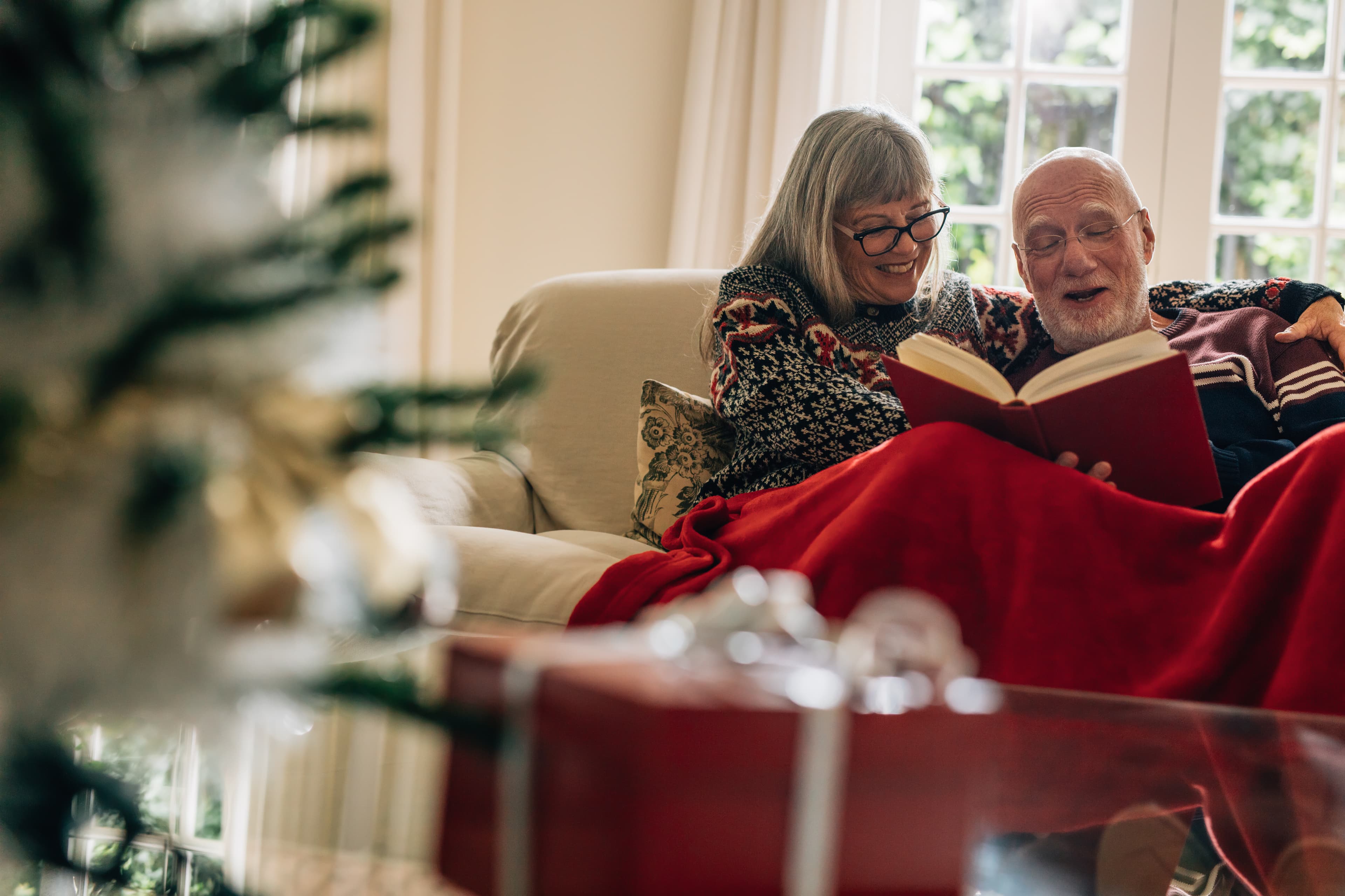Senior couple sitting on a sofa enjoying reading a book with a gift box in the foreground. Smiling couple spending time together reading a book covering themselves in a blanket at home. christmas-traditions-with-books-iceland