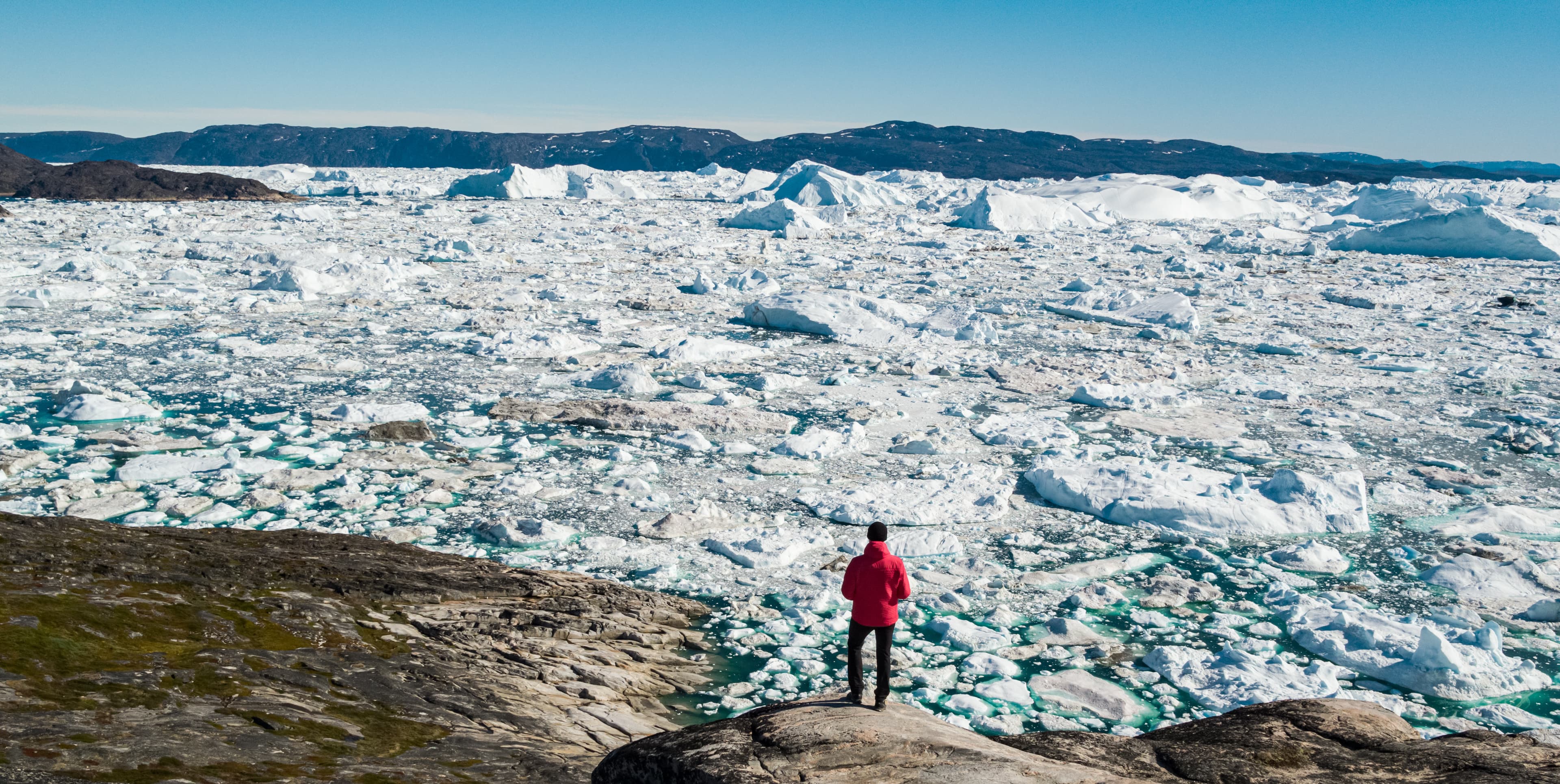 Travel in arctic landscape nature with icebergs - Greenland tourist man explorer - tourist person looking at amazing view of Greenland icefjord - aerial drone image. Man by ice and iceberg, Ilulissat. Travel in arctic landscape nature with icebergs - Greenland tourist man explorer