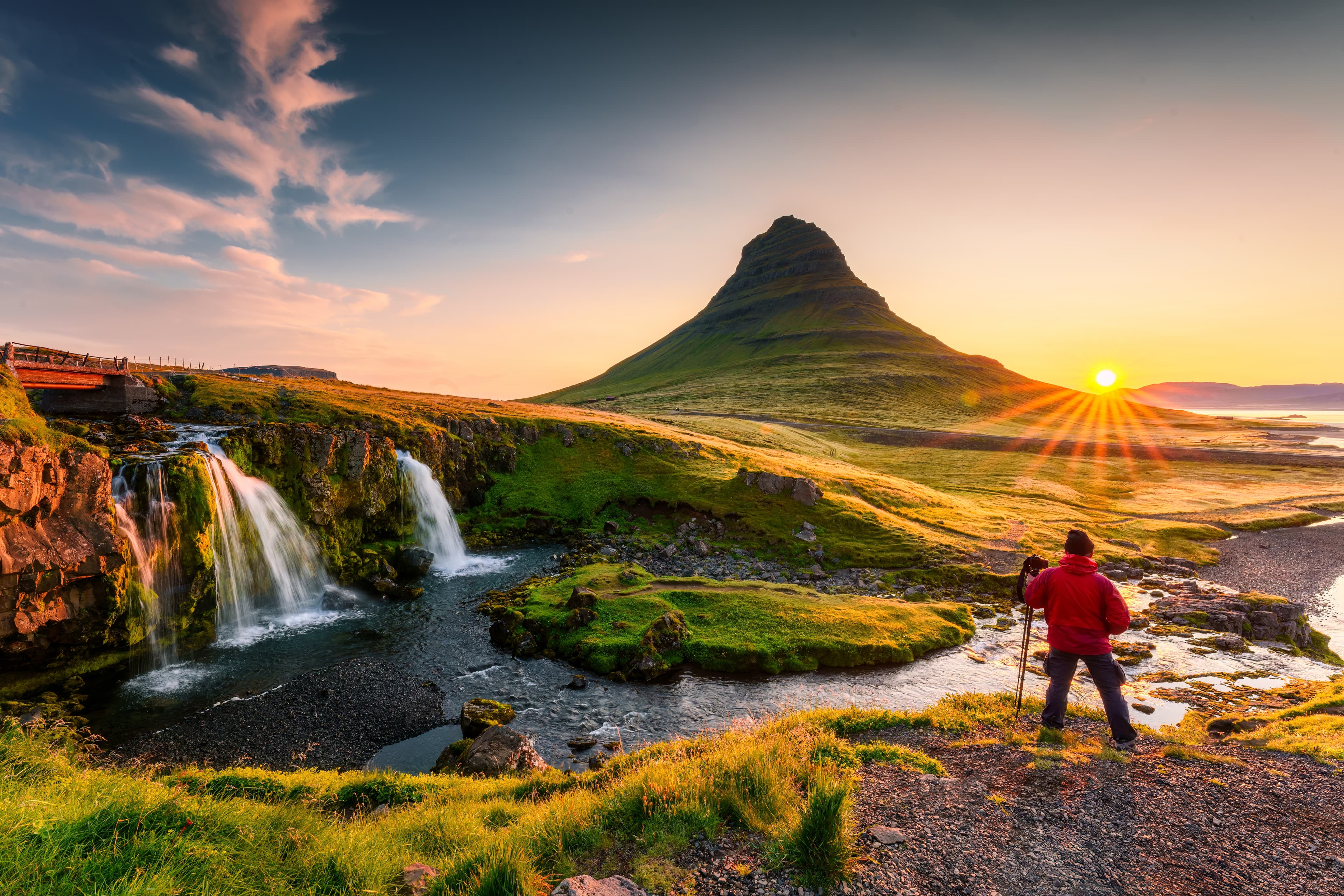 Panorama landscape of sunrise over volcanic Kirkjufell mountain with Kirkjufellsfoss waterfall and photographer man standing in summer at Snaefellsnes peninsula, Iceland Sunrise over volcanic Kirkjufell mountain and photographer man standing in summer at Icelandnaefellsnes peninsula, Iceland