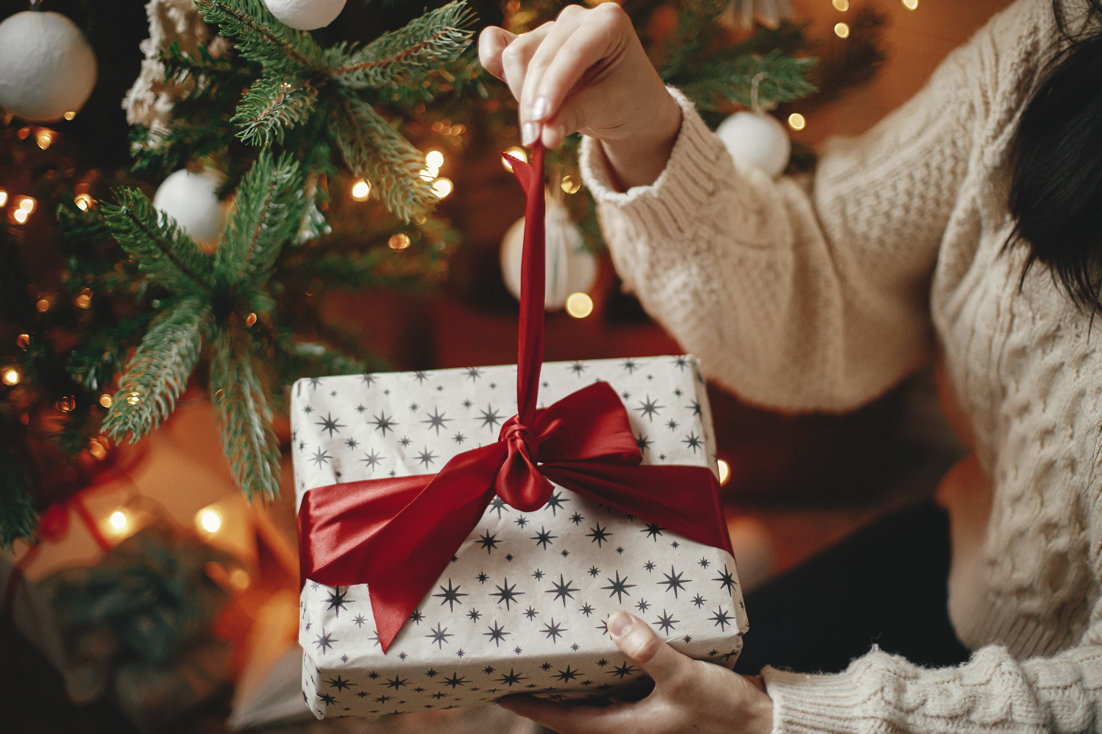 Hands in cozy sweater opening christmas gift with red bow on background of christmas tree with lights. Stylish female holding present with red ribbon in festive room close up. Merry Christmas! Hands in cozy sweater opening christmas gift with red bow on background of christmas tree with lights. Stylish female holding present with red ribbon in festive room close up. Merry Christmas!
