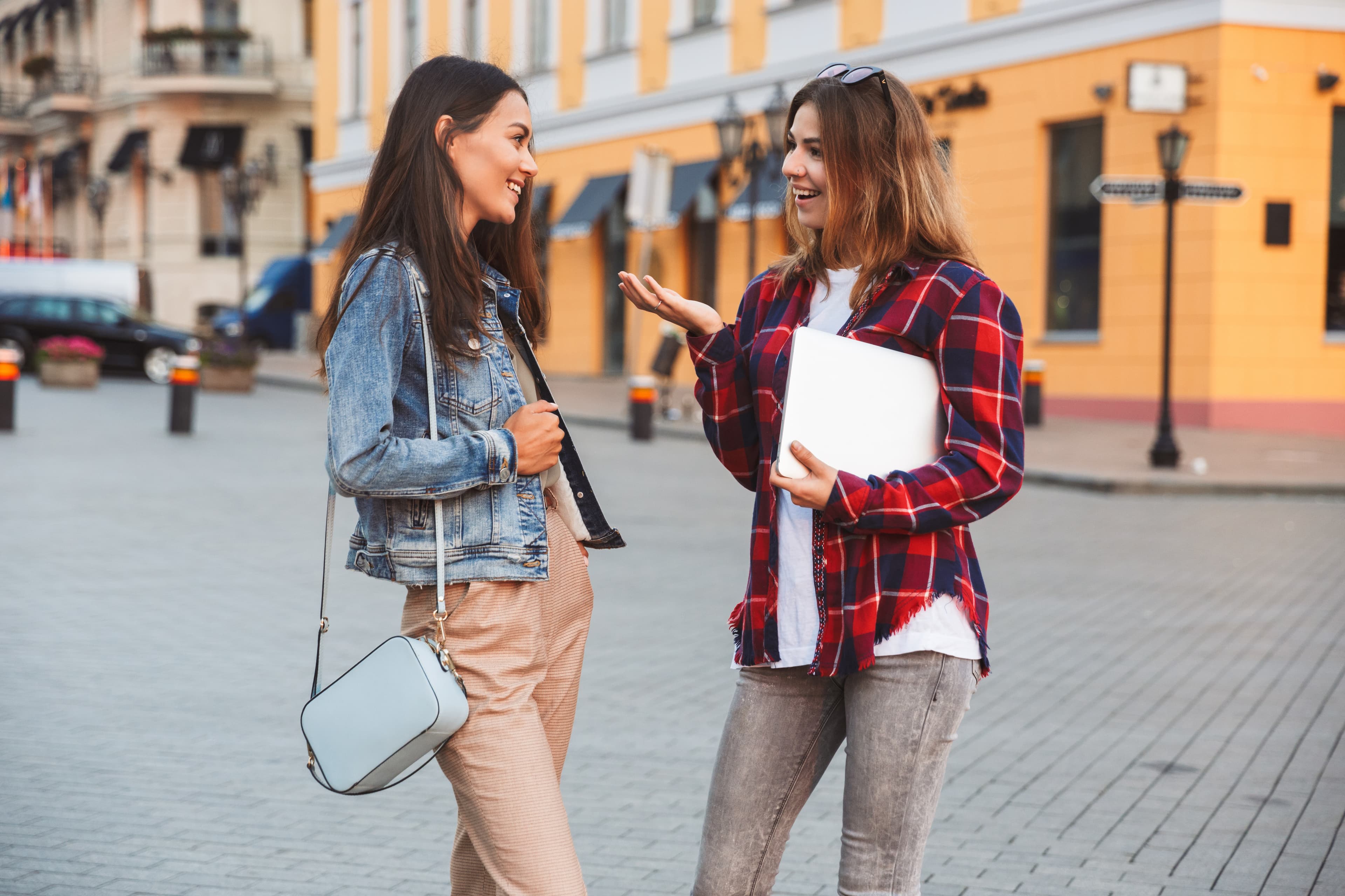Two cheerful young girls friends standing together at the city street, holding laptop, talking Two cheerful young girls friends together
