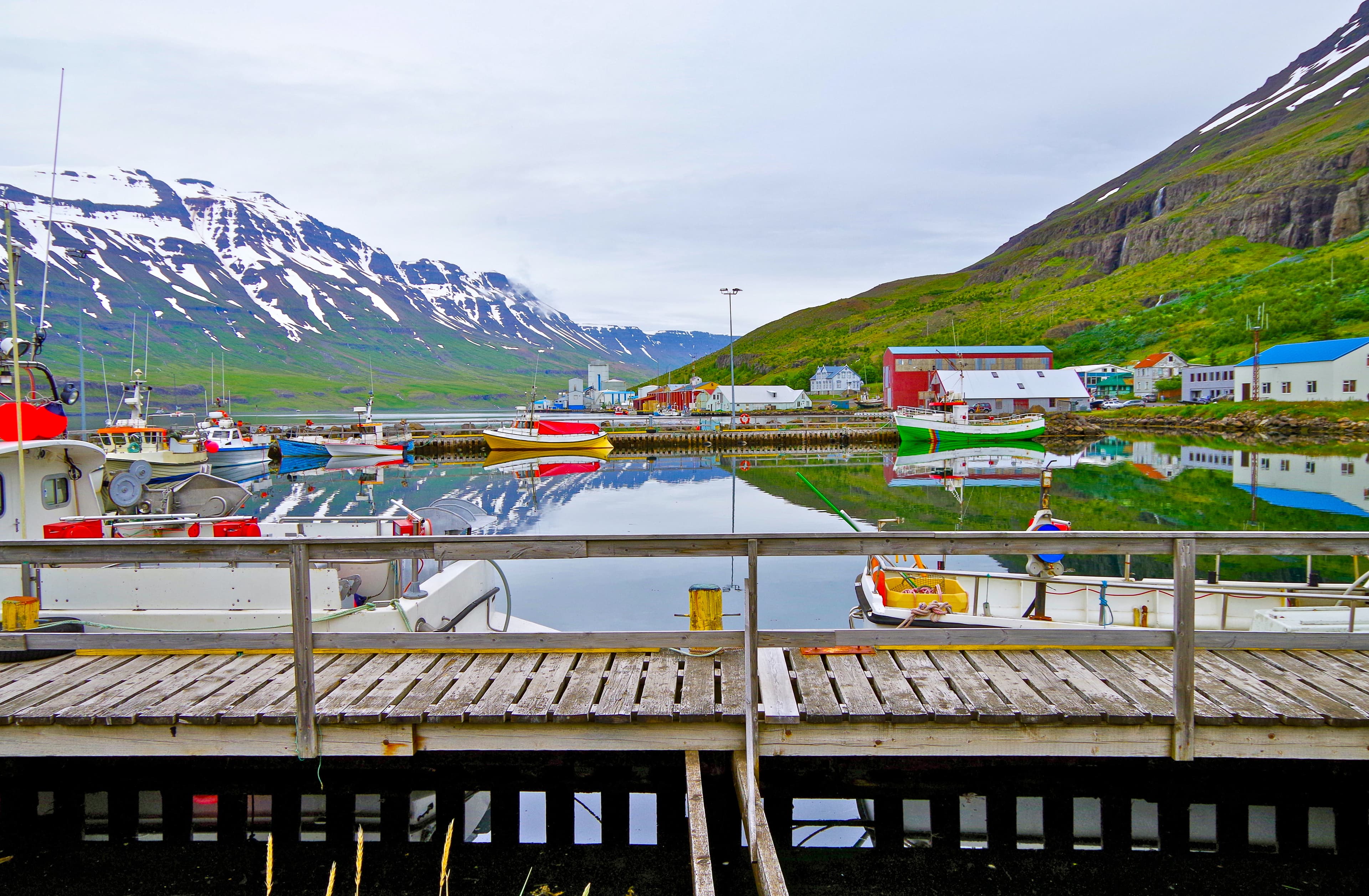Colorful fishing boats and surrounding scenery landscape reflect in even surface of lake or bay in Seydisfjordur Iceland with beautiful mountain range and snow tops with peaceful and tranquil nature