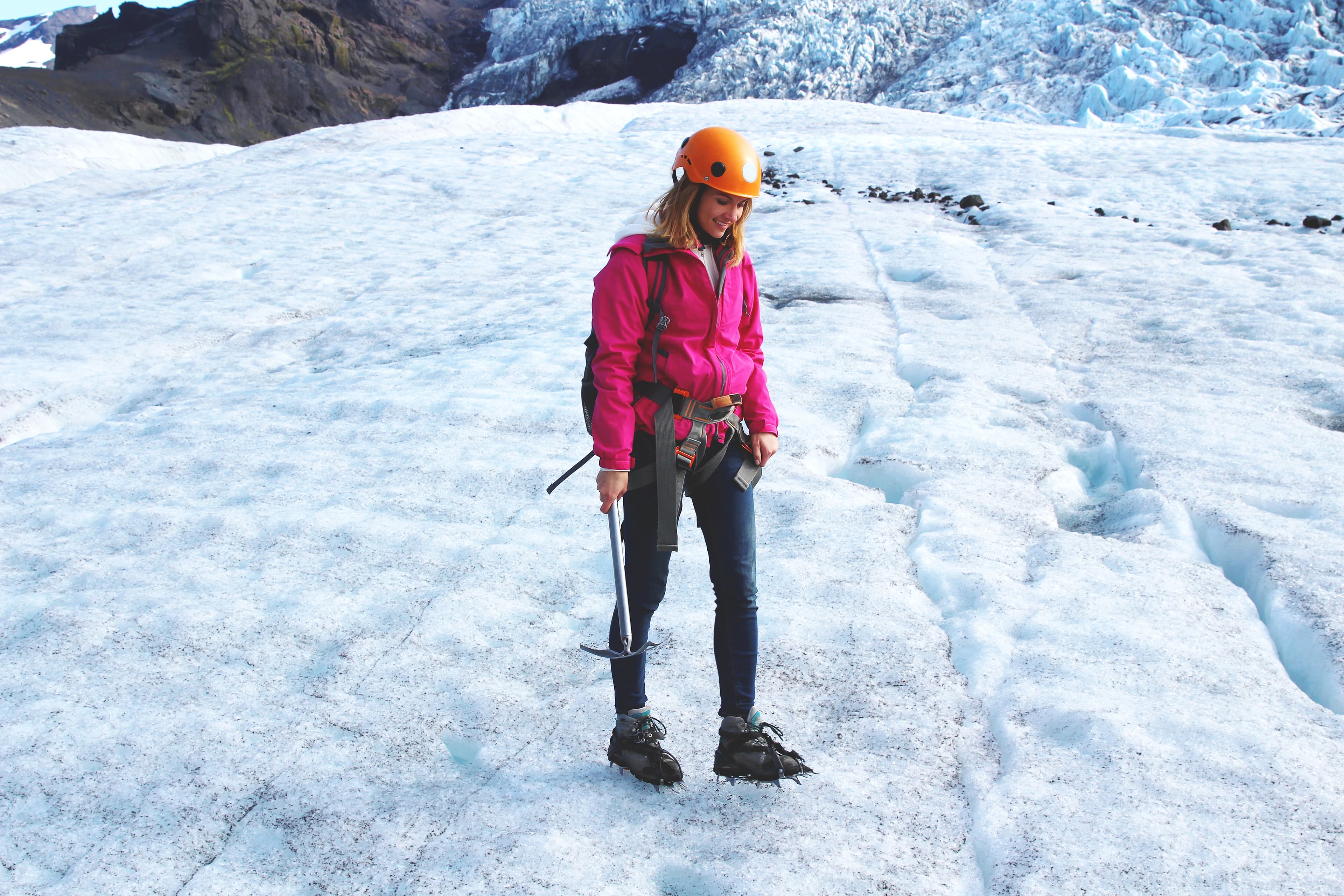Woman wears pink jacket, crampons and helmet, she stands on the glacier in Iceland and holds axe