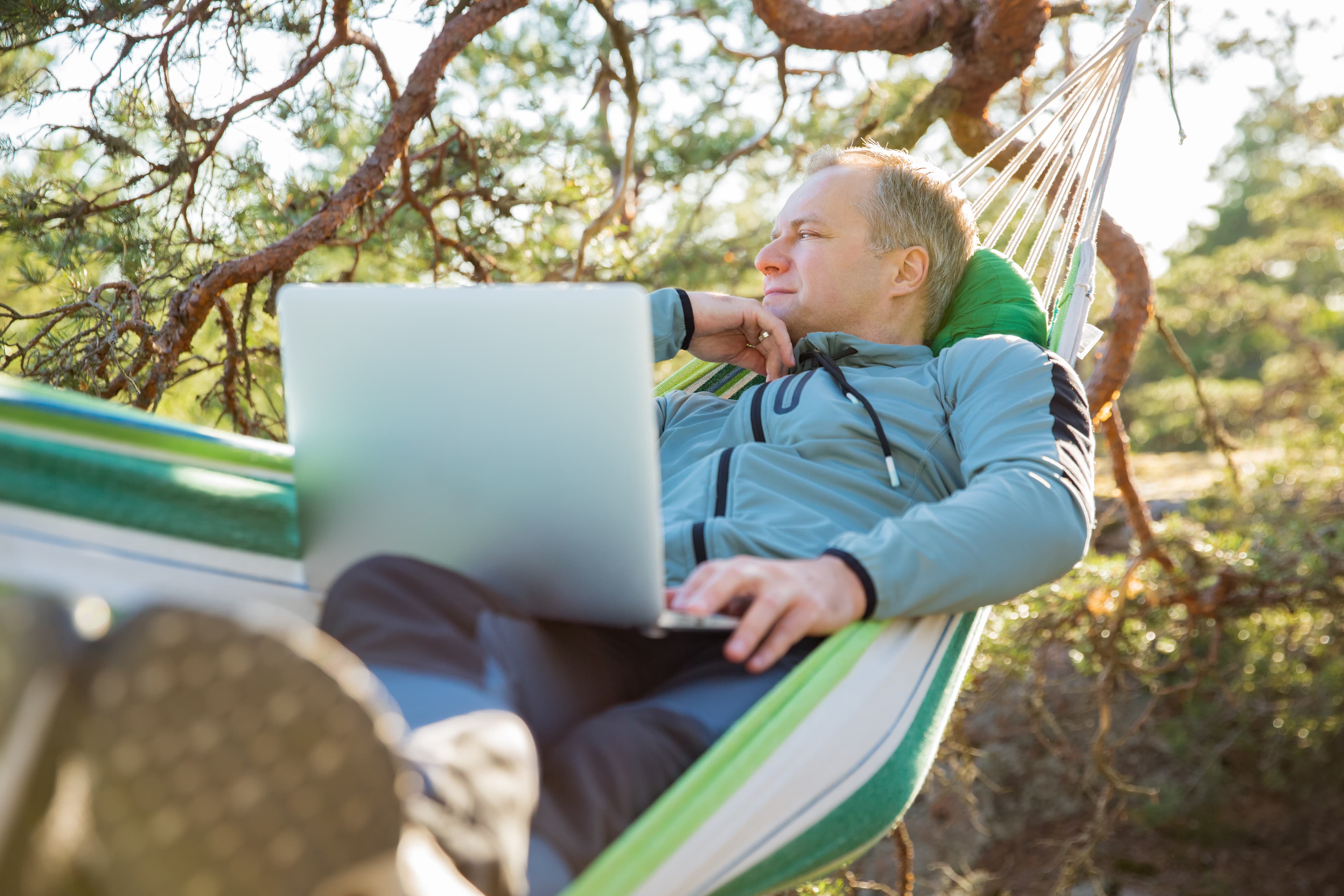 A man working on a laptop while lying in a hammock in the woods. Self-isolation, freelancing, remote work and distancing. Top view of Scandinavian landscape