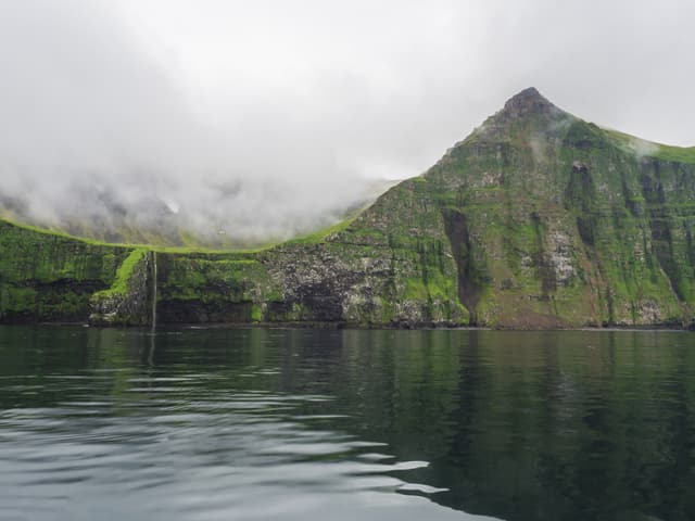 view from boat on steep green Hornbjarg cliffs biggest bird cliffs in Europe, west fjords, remote nature reserve Hornstrandir in Iceland, misty fog ocean and moody sky steep green Hornbjarg cliffs and waterfall, biggest bird cliffs in Europe, west fjords, remote nature reserve Hornstrandir in Iceland, misty fog ocean and moody sky
