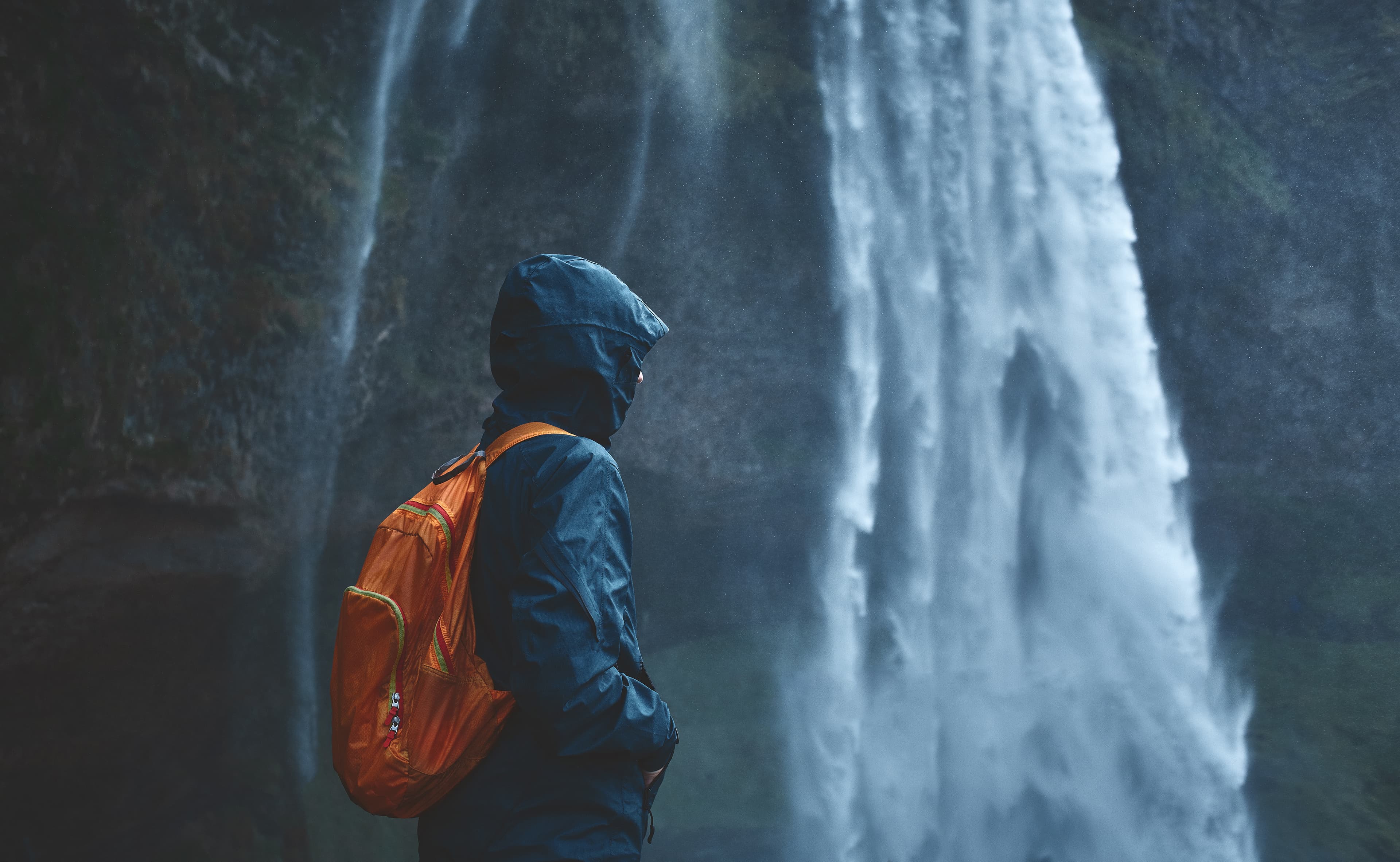 Girl in waterproof clothing stands under the Seljalandsfoss waterfall in Iceland. back view, woman with small orange backpack Seljalandsfoss-waterfall-woman-jacket