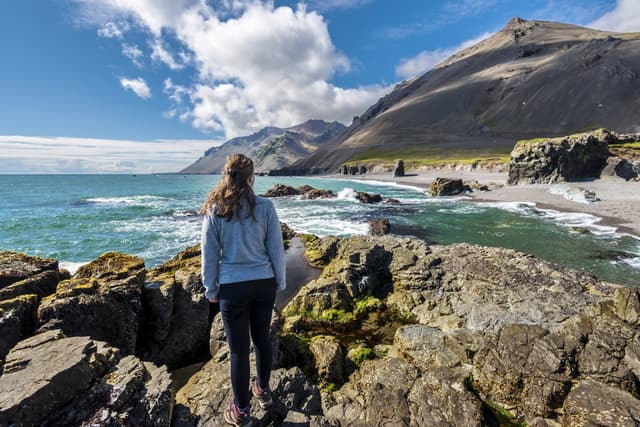Girl teenager is staying on the stone facing to Fauskasandur beach in the eastern Iceland. Maelifell hill is at right background. stone-facing-fauskasandur-beach-eastern-iceland
