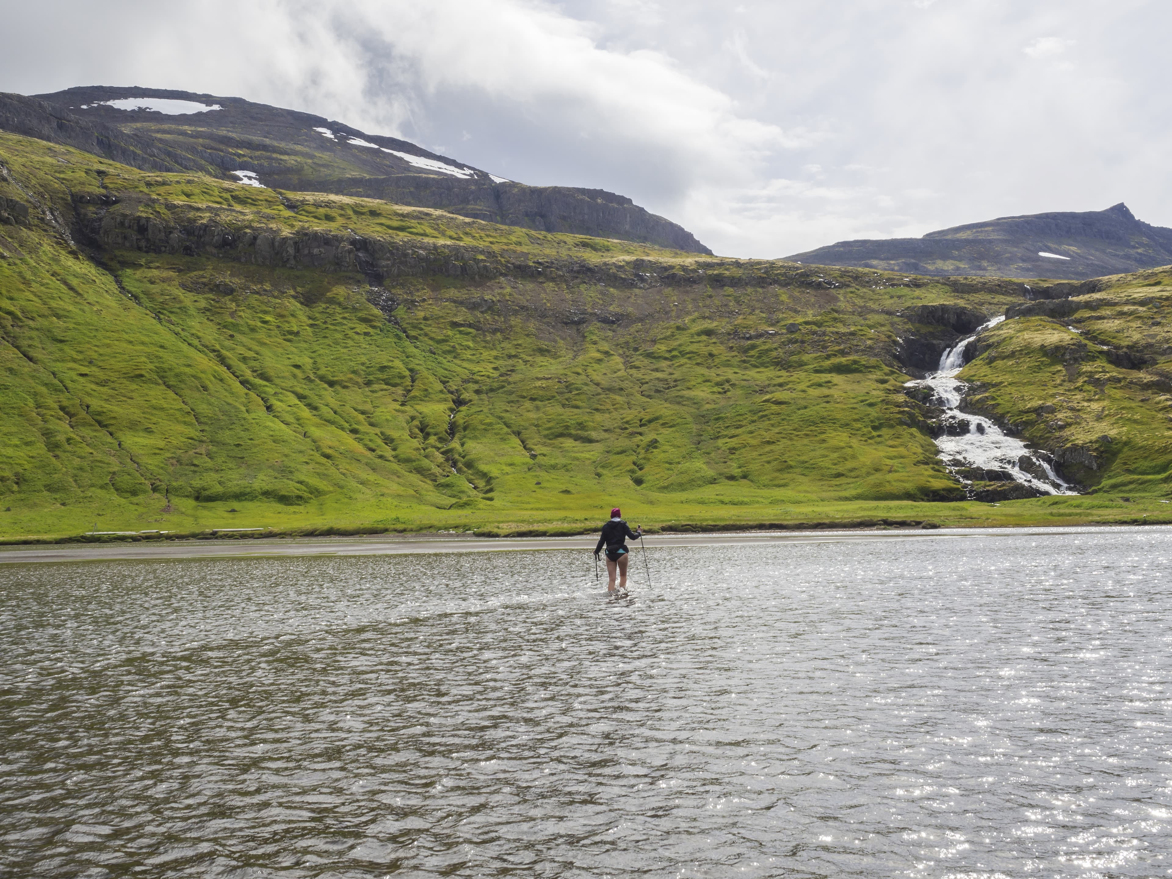 OLYMPUS DIGITAL CAMERA Hiker woman with trekking poles in jacket, purple hat and bare legs crossing river in Iceland Hornstrandir on trek to Hornbjarg cliff, waterfall and green hills in background. Hiking leisure theme