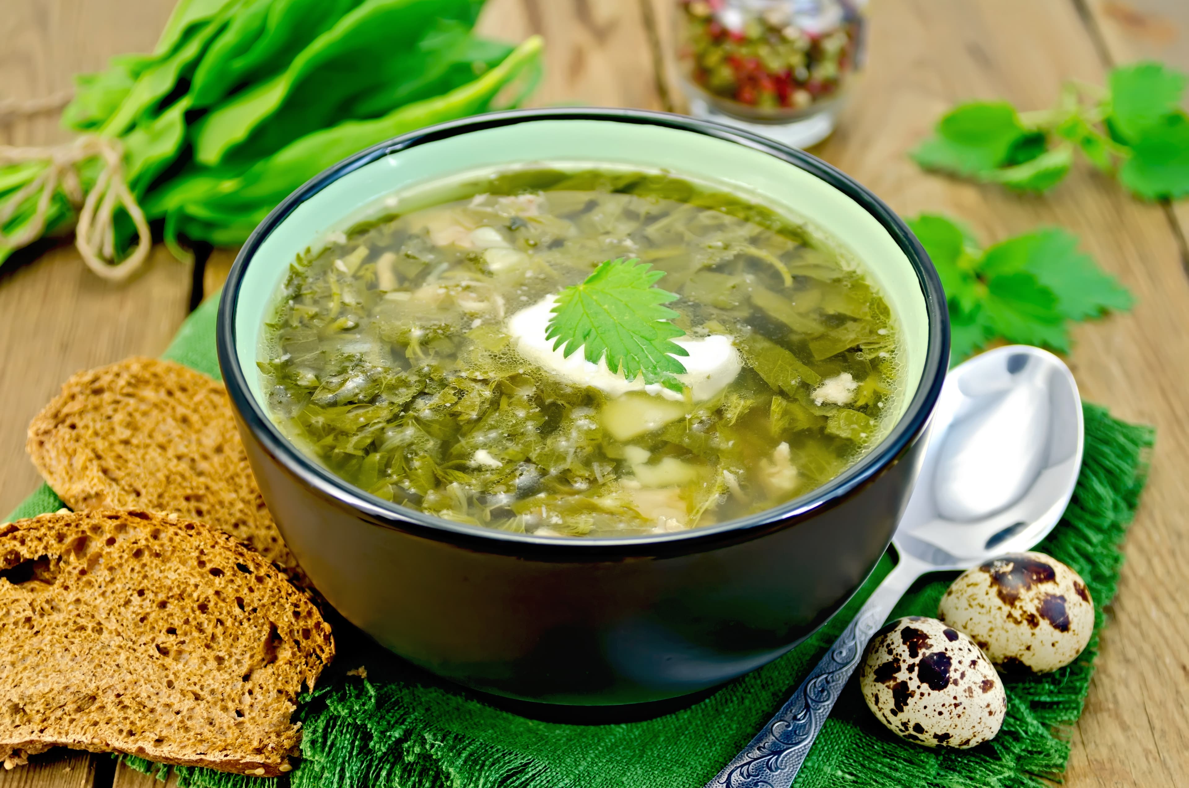 Green soup of sorrel, nettle and spinach in a bowl, spoon, bread, pepper, quail eggs on a wooden board Soup green sorrel and nettles with a spoon on board
