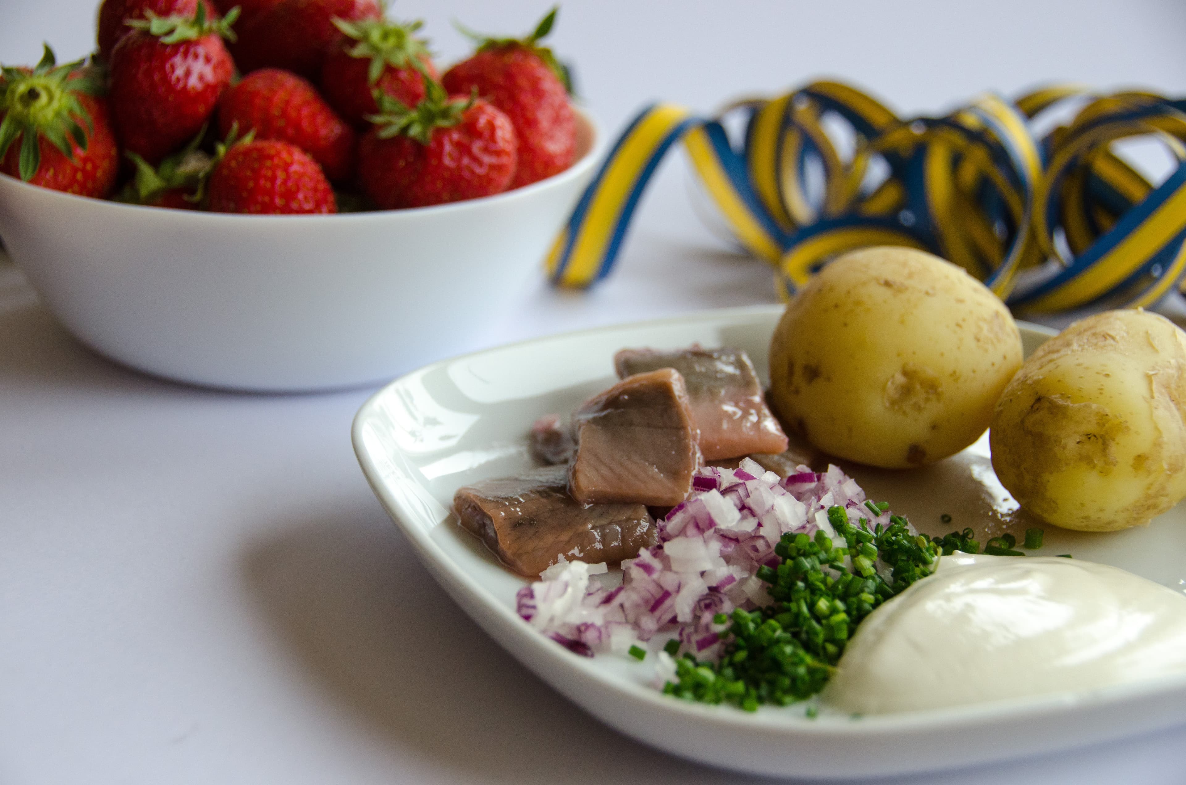 Pickled herring with new potatoes, chives, red onion and sourcream on a porcelain plate. A bowl of strawberries on the side. Sill, färskpotatis och jordgubbar. Herring, new potatoes and strawberries.