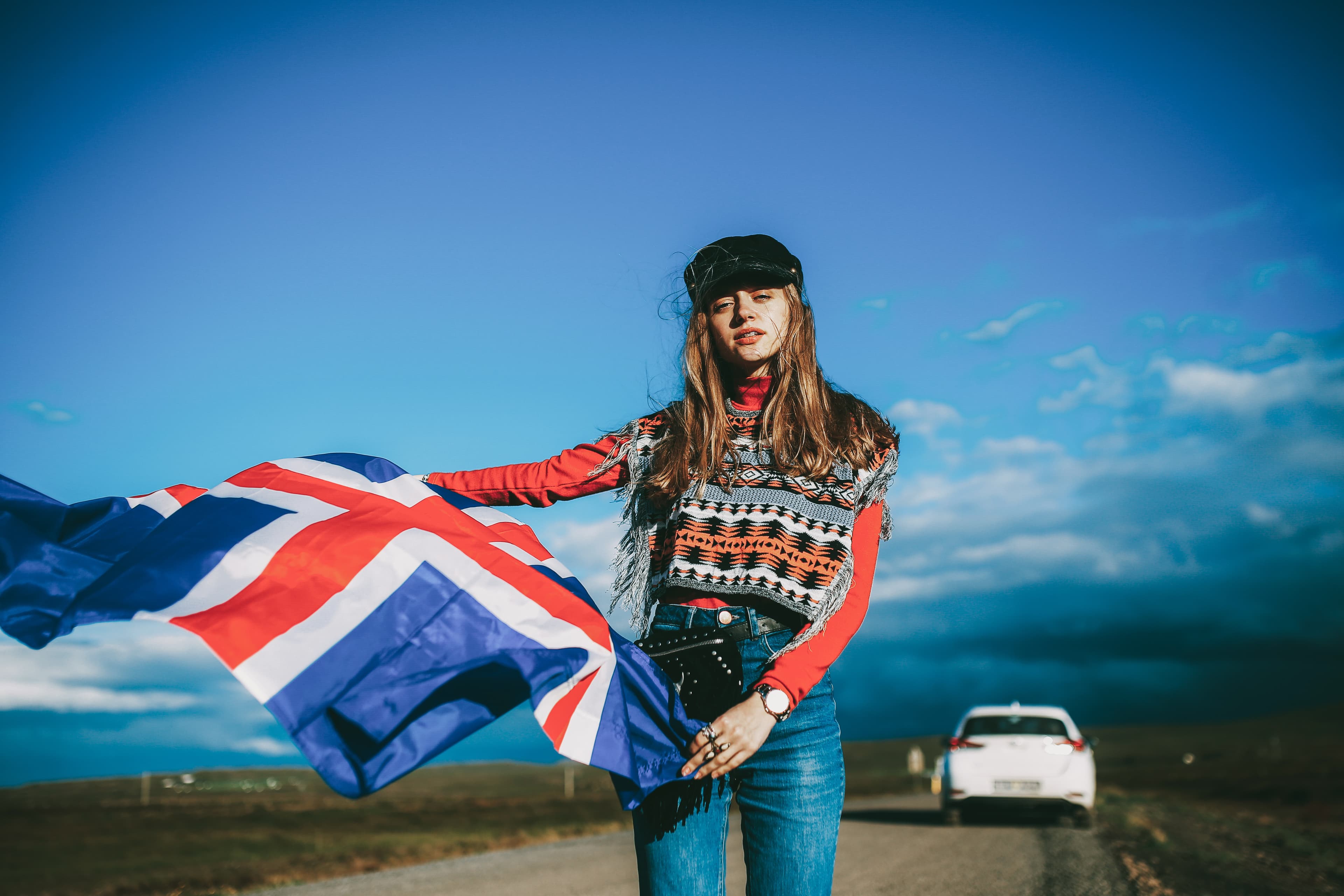 Girl in a colorful cape with the Iceland flag in hands staying on the roadside with the white car at background. Sunny bright colors