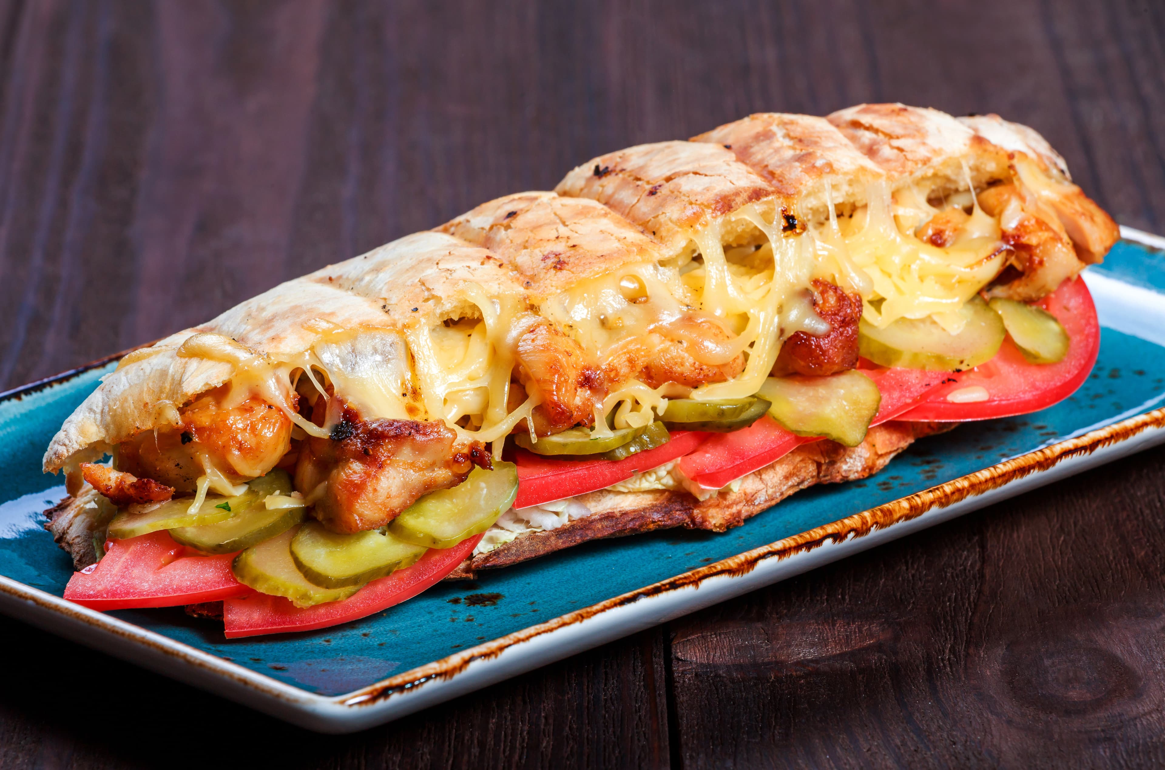 Sandwich from fresh pita bread with fillet grilled chicken, lettuce, slices of fresh tomatoes, pickles and cheese on dark wooden background. Shashlik or Shish kebab. Healthy lunch Sandwich from fresh pita bread with fillet grilled chicken, lettuce, slices of fresh tomatoes, pickles and cheese on dark wooden background. Shashlik or Shish kebab. Healthy lunch