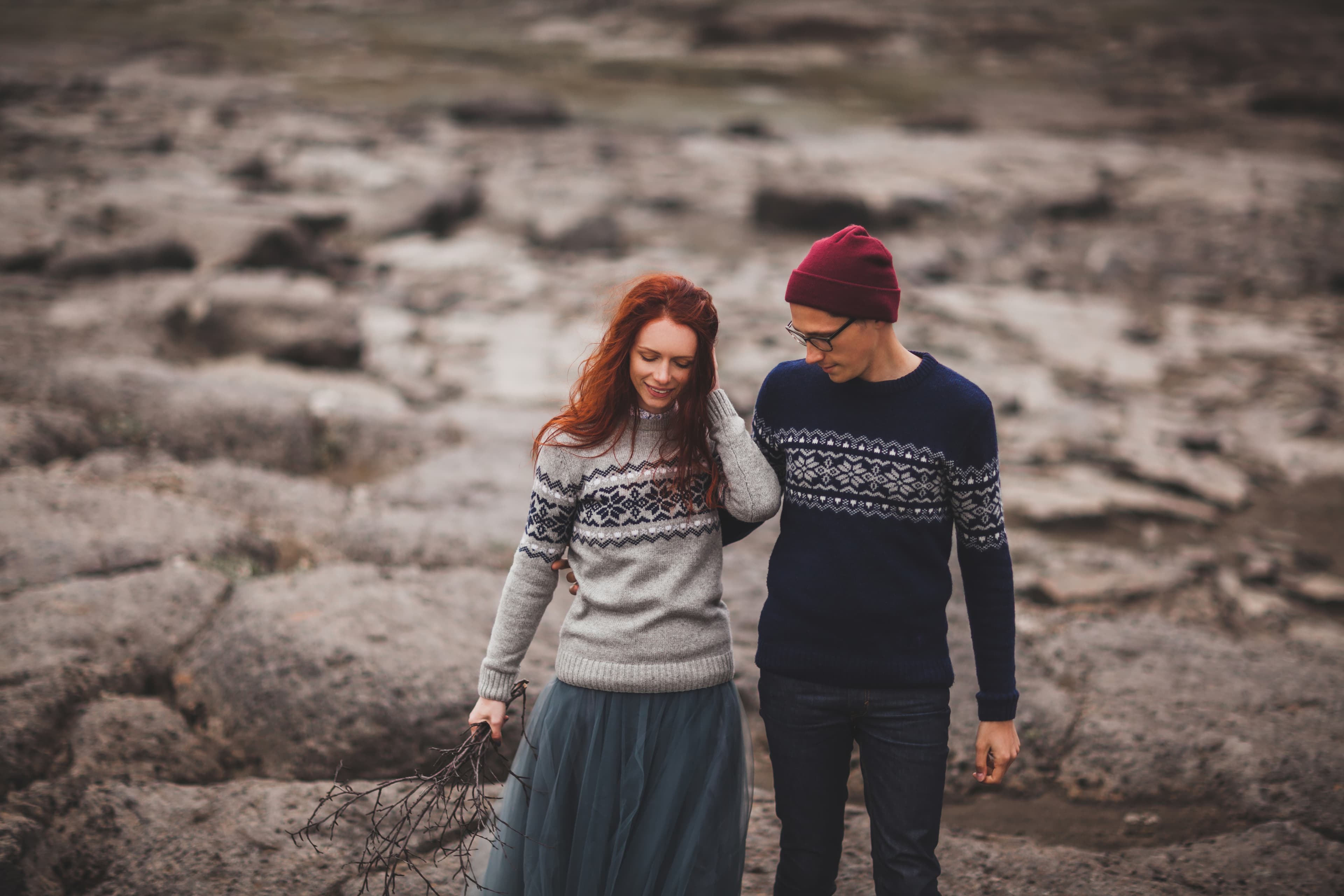 Couple in love walking in Iceland. Stone volcanic desert around, in the middle of nowhere. Dramatic nordic landscape, cold weather. Wearing in traditional wool sweaters, jeans and gray skirt. Couple in love in sweaters walking in Iceland. Stone volcanic desert around
