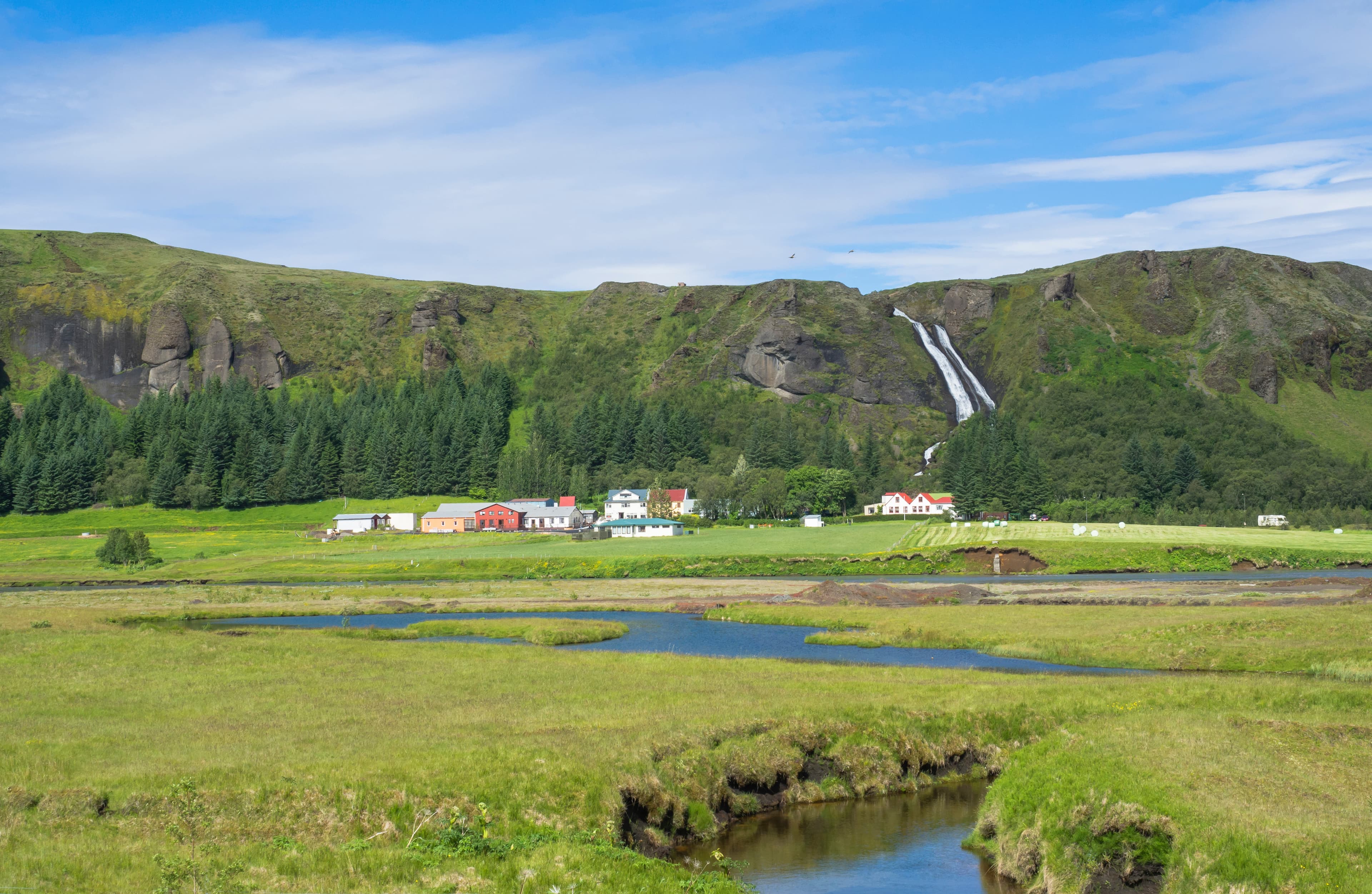 rural landscape with colorful farm houses, blue river stream on green grass meadow with spruce tree forest, hills and beatuful waterfall Systrafoss in Kirkjubæjarklaustur, Iceland rural landscape with colorful farm houses, blue river stream on green grass meadow with spruce tree forest, hills and beatuful waterfall Systrafoss in Kirkjubæjarklaustur, Iceland