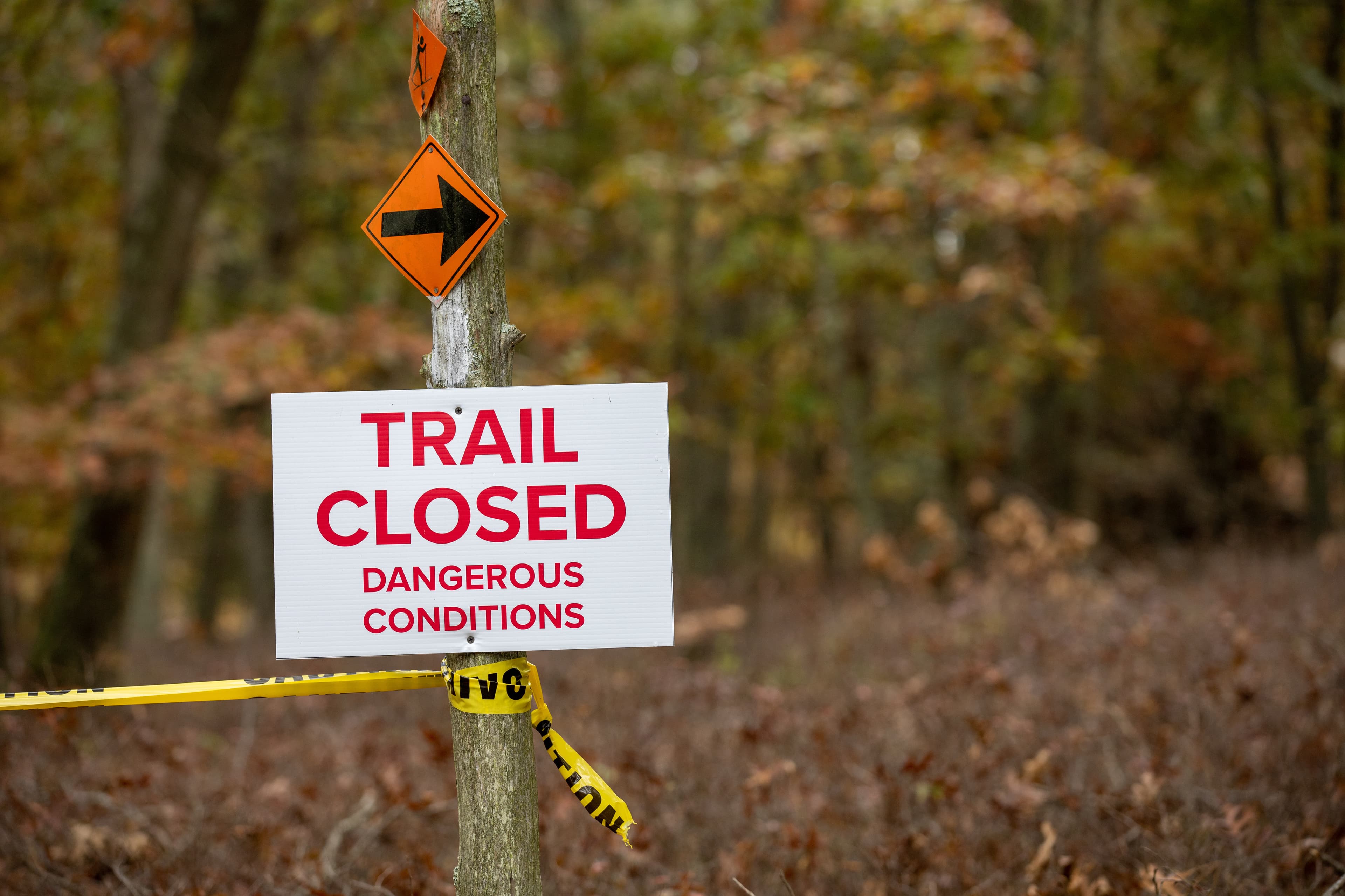 A hiking trail closed due to dangerous conditions. A hazard warning sign and caution tape block the trailhead