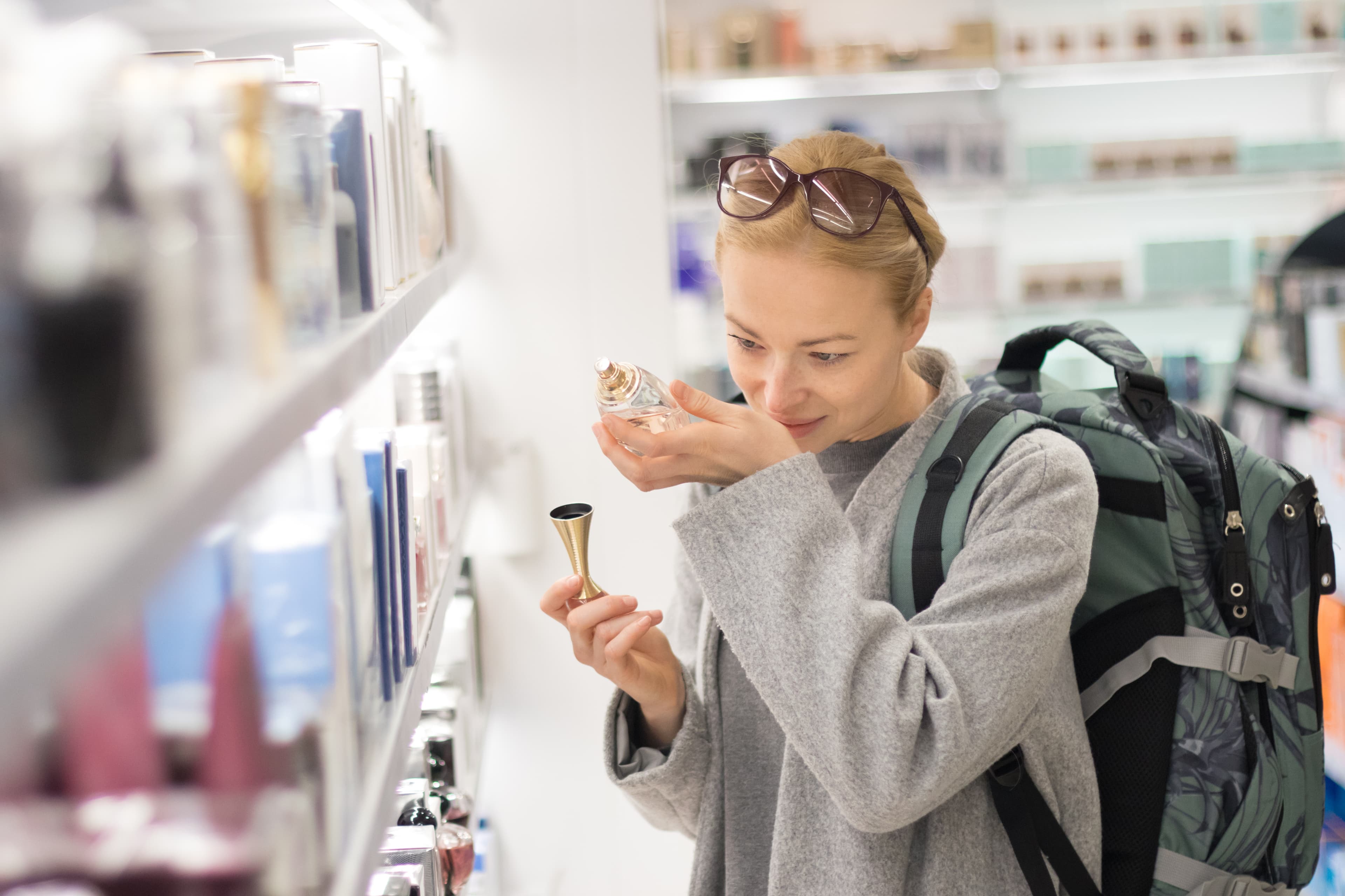 Blond young female traveler wearing coat and travel backpack choosing perfume in airport duty free store. Casual lady testing and buying cosmetics on the go in a beauty store. Blond young female traveler wearing travel backpack choosing perfume in airport duty free store.