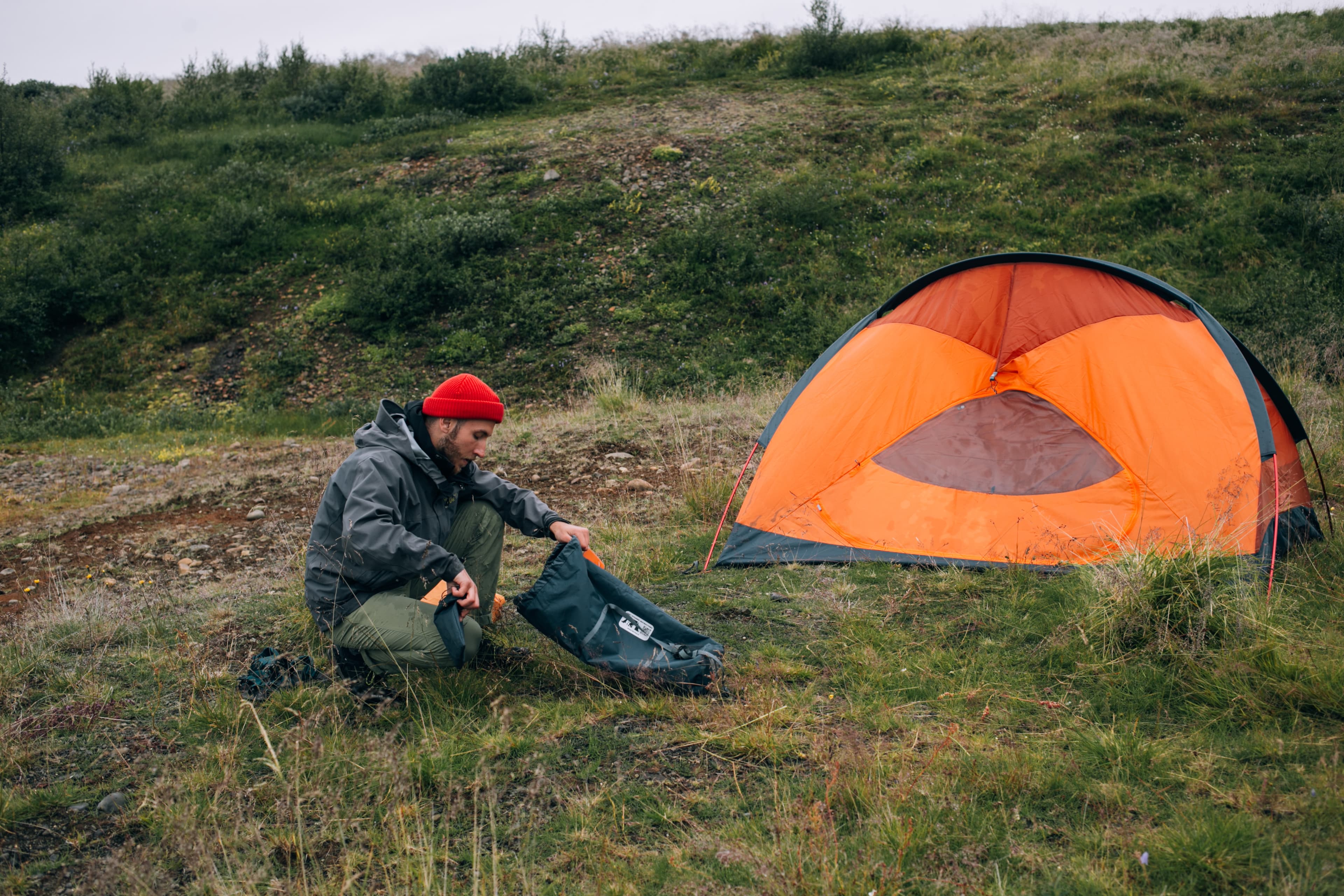 Young male explorer tourist unpacks his dry bag or backpack with camping gear, sleeping matress and hiking food, sets up camp next to tent in wild national park forest or field. Adventure lifestyle Hiker or camper sets up tent in epic scenery